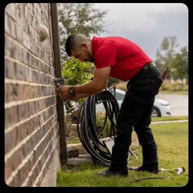 A plumber in a red shirt services an outdoor water spigot on the side of a home