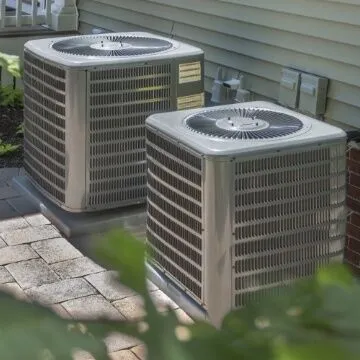 Two large air conditioning units are installed outdoors beside a house, positioned on a paved surface near the wall. The units are surrounded by greenery, highlighting the outdoor setting.