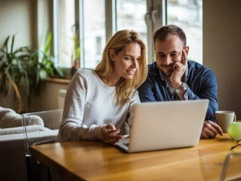 A couple is seated at a table, intently looking at a laptop screen while sharing a moment of engagement. The woman appears excited, pointing at the screen, while the man listens attentively, resting his chin on his hand, suggesting a collaborative or enjoyable activity.