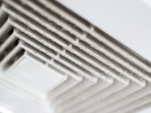 A close-up view of an air conditioning vent with visible dust and debris accumulation on the slats. The vent appears to be part of a ceiling-mounted unit, highlighting the need for maintenance to ensure proper airflow.