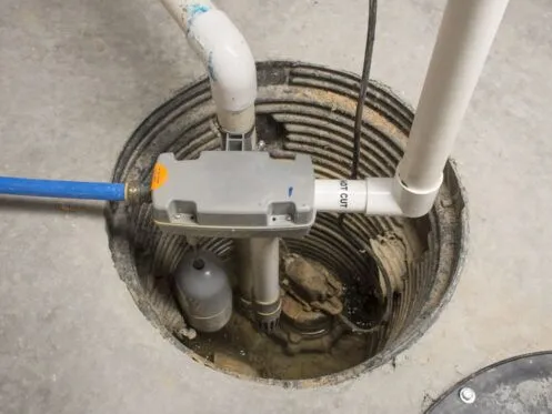 A sump pump installed in a concrete basement floor with PVC piping connected to the system. The pit appears partially filled with water and debris.