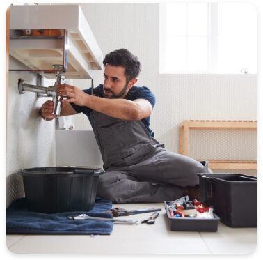 A plumber in gray overalls works on a pipe under a sink, surrounded by tools and a bucket. The scene takes place in a bright bathroom with a window in the background.