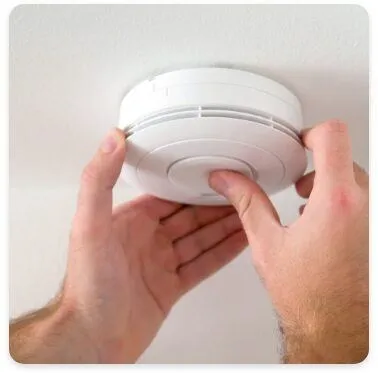 A person is installing or checking a smoke detector mounted on a ceiling. Their hands are positioned on the device, demonstrating an action related to home safety equipment.