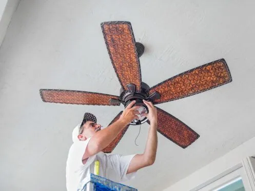 A man standing on a ladder installs or repairs a ceiling fan with large decorative blades. He is focused on wiring the fan while wearing a white shirt and a patterned cap.