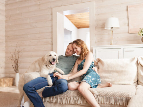 A couple sits happily on a cozy couch with their golden retriever, sharing a warm moment in a bright, minimalist living room. The room features light wood-paneled walls and soft, neutral decor.