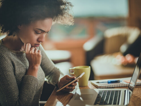 A woman sits at a desk with a thoughtful expression while looking at her smartphone, with a laptop and a mug beside her. The room is softly lit, creating a cozy working environment.