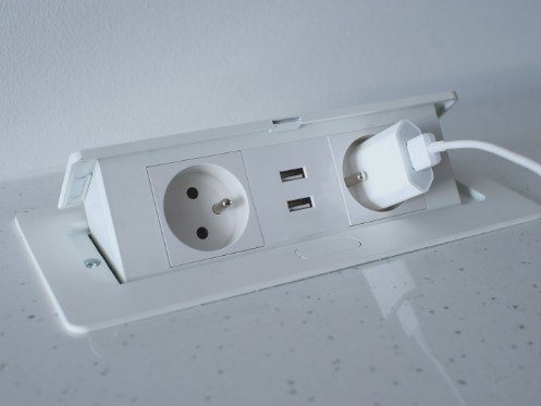 A countertop power outlet is shown with two standard sockets and two USB ports. A charging plug is connected to one of the sockets, while the outlet is set into a sleek surface, complementing a modern aesthetic.