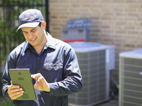 A technician in a navy uniform and cap uses a tablet while standing outdoors near air conditioning units. The brick building and HVAC equipment are visible in the background.