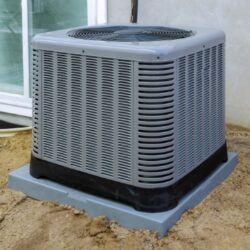 A gray air conditioning unit is placed on a concrete pad in a sandy area, positioned close to a window. The unit features a grille design and a circular top vent for air circulation.