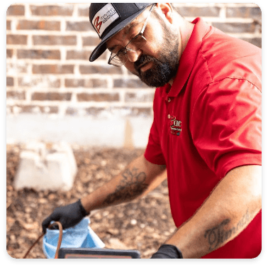 A technician in a red shirt and black gloves, wearing a branded cap, works outdoors with a focused expression while handling equipment. A brick wall is visible in the background.