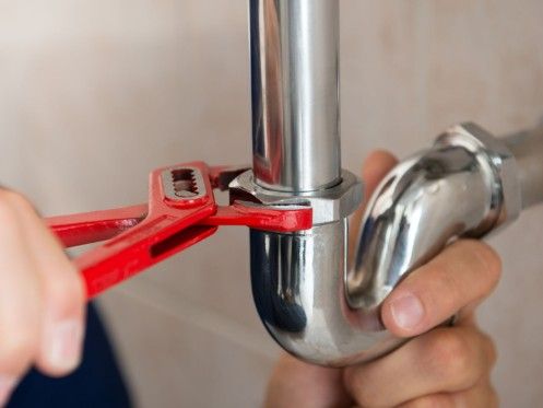 A plumber is using a red adjustable wrench to tighten a metal pipe joint under a sink. The shiny silver pipes and fittings are clearly visible, highlighting the tools and technique used in plumbing work.