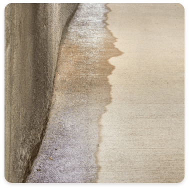 A close-up view of a concrete surface showing a distinct line where wet and dry areas meet. The texture of the concrete wall and floor is visible, indicating water pooling or drainage along the edge.