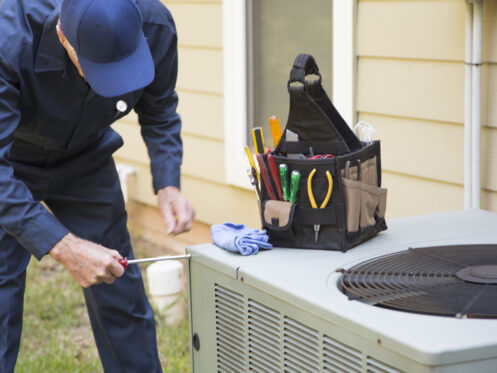 A technician in a navy uniform works on an outdoor air conditioning unit with a screwdriver. A tool bag filled with various tools sits on top of the unit.