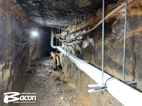 A view of an underground tunnel with exposed pipes running along the walls and ceiling, supported by metal braces. The Bacon Plumbing Heating Air Electric logo is visible in the bottom left corner.