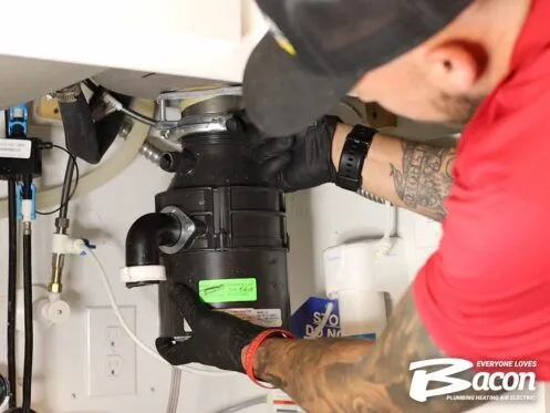 A technician in a red shirt and black gloves installs or repairs a garbage disposal under a kitchen sink. The Bacon Plumbing Heating Air Electric logo is visible in the lower right corner.