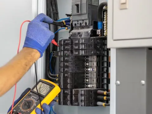 A technician wearing blue gloves uses a multimeter to test an electrical panel with exposed wiring. The device displays a reading, and several circuit breakers are visible.