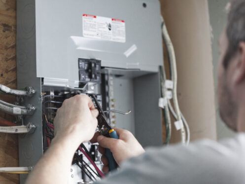 A person is working on an electrical panel, using tools to connect wires while looking intently at the panel. The image focuses on the individual's hands and the panel, highlighting the intricate wiring.