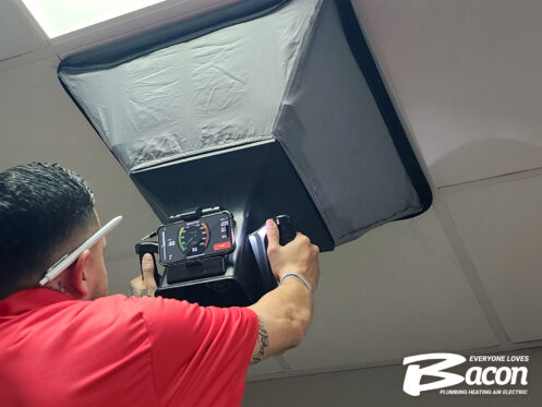 A technician in a red shirt holds a device under a ceiling vent with a large airflow testing hood attached. The Bacon Plumbing Heating Air Electric logo appears in the bottom right corner.