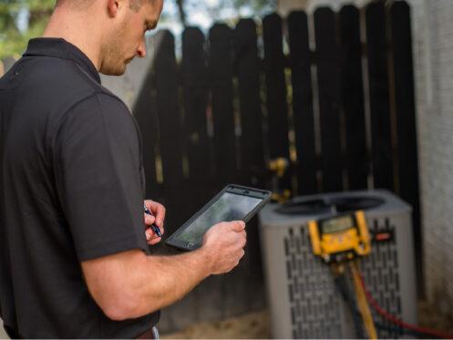 A technician stands outside, holding a tablet while checking the readings from an air conditioning unit. The outdoor unit is connected to hoses, and a wooden fence serves as the backdrop.