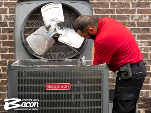 A technician in a red shirt works on an outdoor Goodman air conditioning unit with its fan cover removed. The Bacon Plumbing Heating Air Electric logo is visible in the lower left corner.