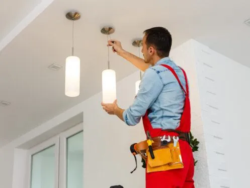 A technician in red overalls is replacing a light fixture, reaching up to attach a pendant light from the ceiling. The work area is bright and modern, featuring multiple hanging lights.