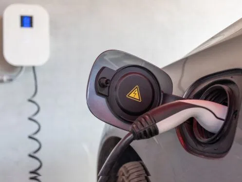 An electric vehicle is connected to a charging station, with the charging plug inserted into the vehicle's charging port. The charging station is visible in the background, indicating the vehicle is actively charging.