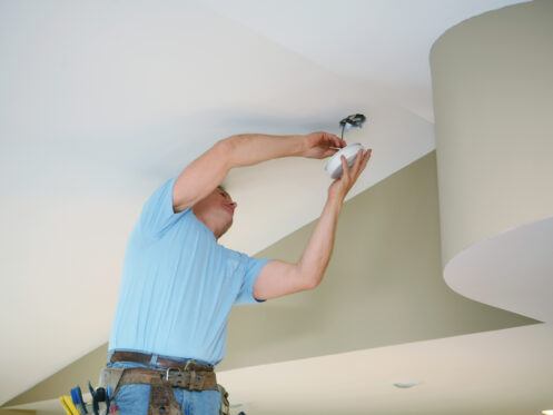 A technician is installing a ceiling light fixture while standing on a ladder. He is focused on connecting the wiring inside a circular ceiling mount, wearing a light blue shirt and a tool belt.