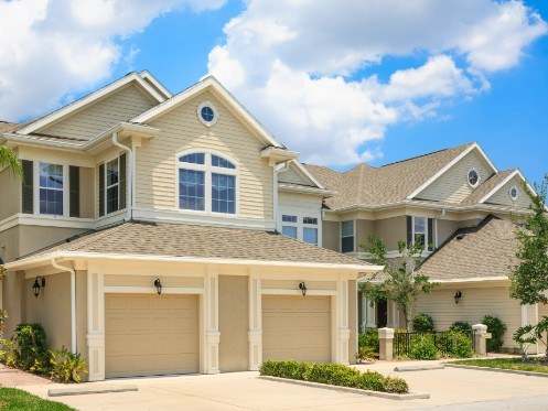 The image showcases a charming two-story house with light beige siding and a classic design, featuring a symmetrical layout. Two garage doors are visible at the front, surrounded by well-maintained landscaping under a bright blue sky dotted with fluffy clouds.