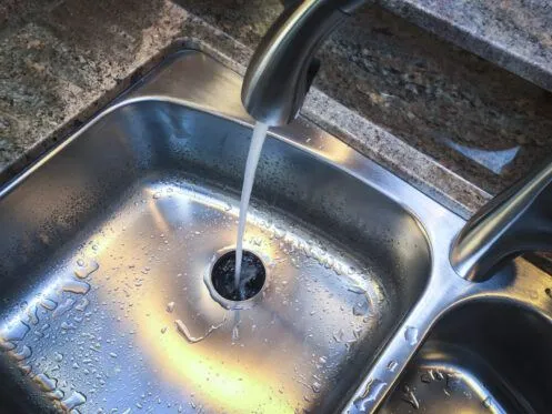 Water is flowing from a kitchen faucet into a stainless steel sink, creating ripples on the surface. The sink features a drain at the center, surrounded by a wet and shiny surface.