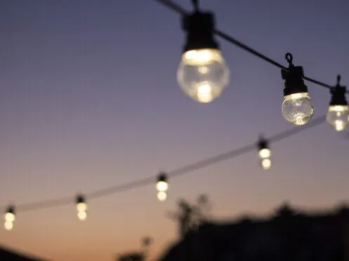 String lights glow against a soft sunset backdrop, creating a warm and inviting ambiance. The lights are evenly spaced, with the sky transitioning from light to dark hues, enhancing the evening's atmosphere.