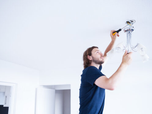 A man in a navy shirt installs or repairs a chandelier on a white ceiling, using a screwdriver to adjust the fixture. The room is brightly lit with white walls and doors.