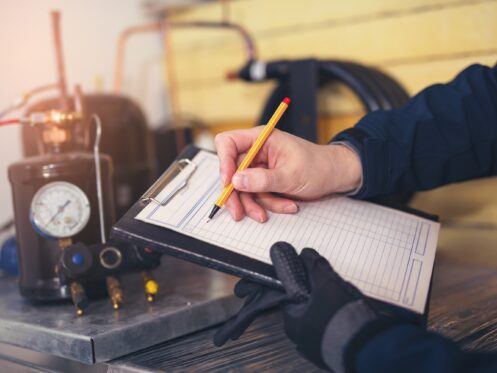 A technician holds a clipboard with a checklist while preparing to record information. In the background, equipment and tools, including a pressure gauge, are visible on a workbench.