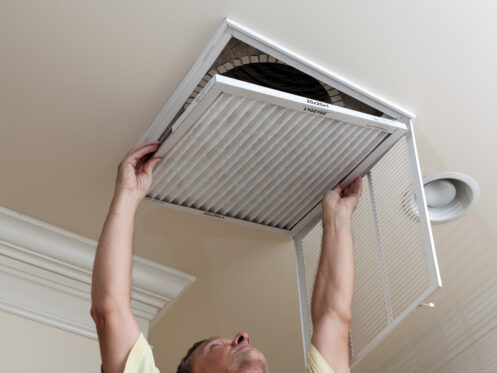 A person replaces an air filter in a ceiling vent, with the vent cover opened and the old filter partially removed. The ceiling has crown molding and a nearby recessed light fixture.