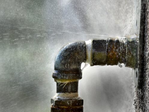 Water is spraying out from a leaky metal pipe joint, with droplets visibly splashing in all directions. The close-up image highlights the rusty texture of the pipe fittings and the force of the escaping water.