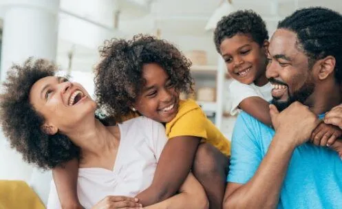 A joyful family of four shares a playful moment, with the parents smiling and their two children happily engaging with them. The scene captures a warm and affectionate family interaction in a bright living room.