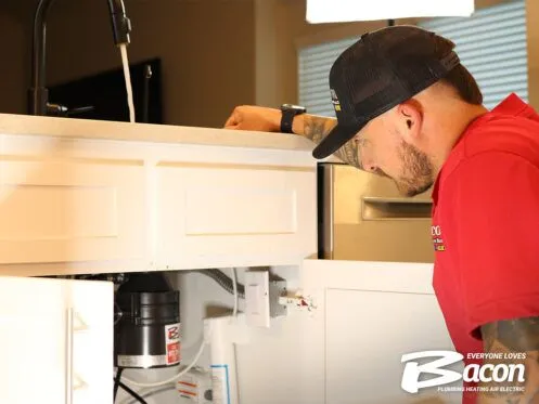 A technician in a red shirt and black cap inspects the plumbing under a kitchen sink with a garbage disposal unit visible. The Bacon Plumbing Heating Air Electric logo is displayed in the bottom right corner.