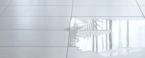 A puddle of water reflects light on a smooth white tiled floor, indicating a potential leak or spill. The reflection suggests the presence of nearby windows or other light sources.