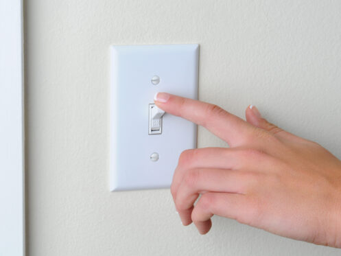 A hand with neatly manicured nails flips a light switch on a plain beige wall. The switch plate is white with two visible screws.