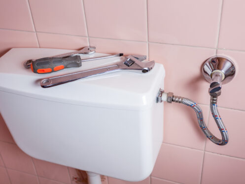 A set of tools, including a wrench and screwdriver, rests on top of a toilet tank connected to a flexible water supply line. The background features pink tiled walls.