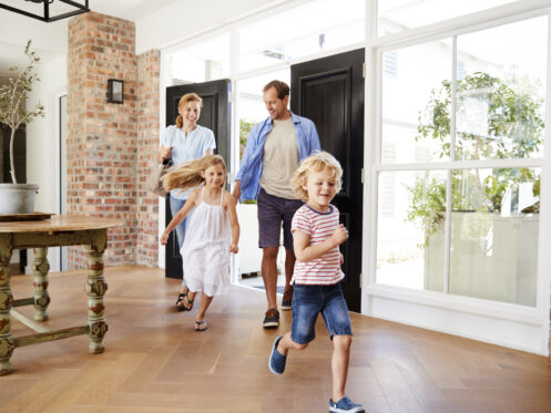 A family of four, including two young children and their parents, happily enters a bright, modern home. The children run ahead, excited, while the parents follow, smiling.