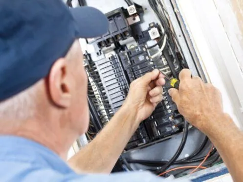 An electrician in a blue cap is working on a breaker panel, using a screwdriver to adjust wiring connections. The panel contains various circuit breakers and electrical wires.