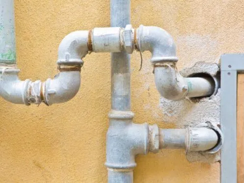 A series of old metal pipes are connected on the exterior of a building, with some pipes entering the wall through visible openings. The pipes show signs of wear and rust, suggesting they may need maintenance or replacement.