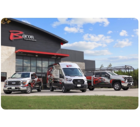 Three branded service vehicles are parked in front of a building with a "Bacon Plumbing Heating Air Electric" logo. The vehicles include a truck, a van, and a pickup with a ladder rack.