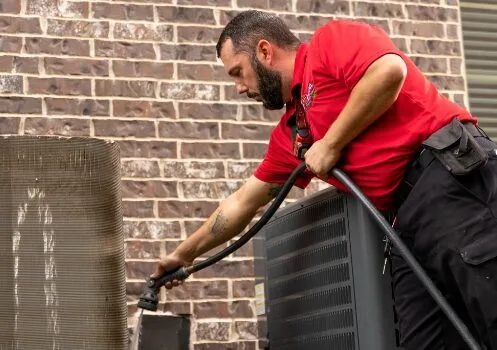 A technician in a red shirt is cleaning an outdoor air conditioning unit with a hose, spraying water on the unit's condenser coil. The scene shows maintenance work being done on the exterior part of the HVAC system.