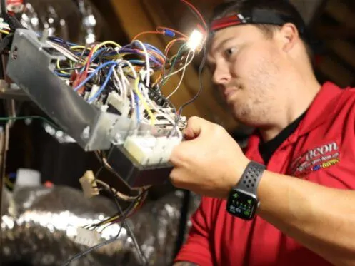 A technician wearing a headlamp is working on a complex network of wires, examining an electrical component in an attic space. The technician is wearing a red shirt and a smartwatch, focusing on the electrical connections.