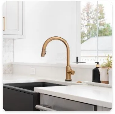 A modern kitchen sink with a sleek gold faucet is installed on a white countertop, next to a black soap dispenser and potted plant. A window in the background allows natural light to brighten the space.