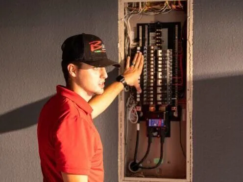 A technician in a red shirt and black cap is examining a breaker panel, pointing to the circuit breakers inside the open electrical box. The technician appears to be explaining or inspecting the wiring and components.