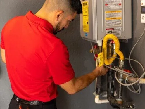 A technician in a red shirt is servicing a tankless water heater, adjusting the pipes and valves connected to the unit. The setup includes various pipes and a yellow gas line.