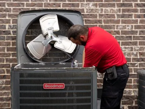 A technician in a red shirt is performing maintenance on a Goodman air conditioning unit, with the fan and internal components exposed. The unit is positioned against a brick wall outdoors.