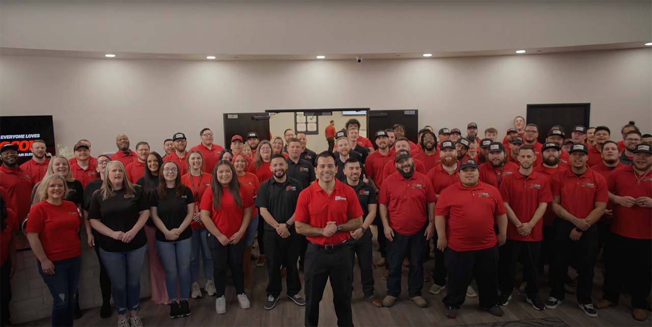 A large group of employees, most wearing red shirts and some in black, are standing together for a group photo indoors. The setting appears to be a company gathering, with the logo "Everyone Loves Bacon" partially visible on a screen in the background.